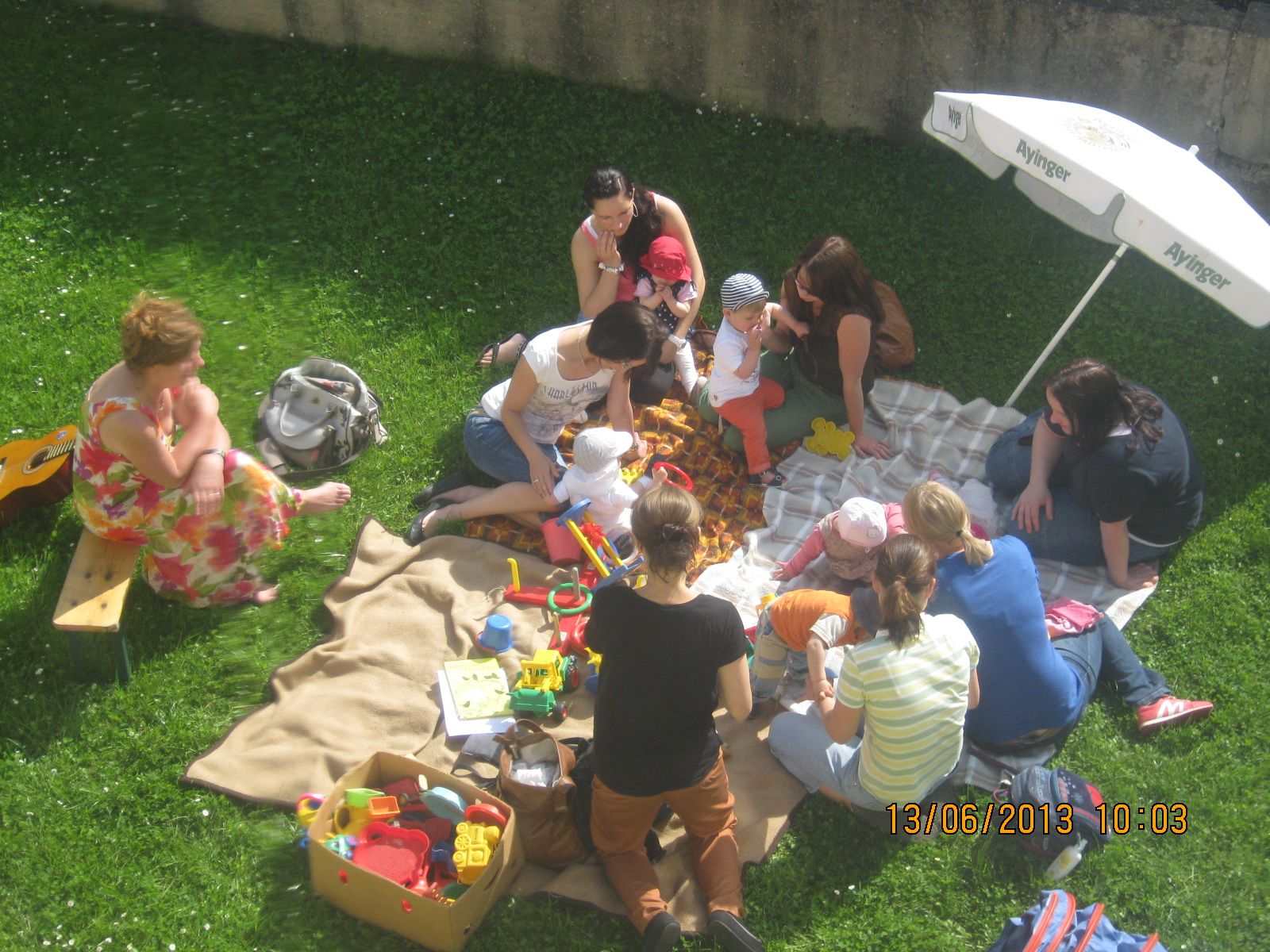 Eine Gruppe von Müttern und Kindern spielt gemeinsam auf einer Decke im Freien, umgeben von Spielzeug.