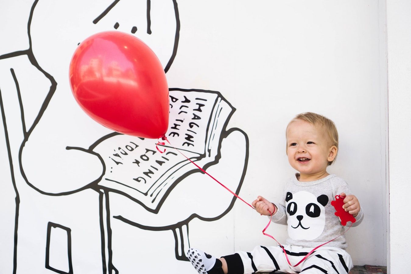 Ein fröhliches Kleinkind sitzt mit einem roten Ballon und einer Spielzeugfigur vor einer Wand mit einer Zeichnung.