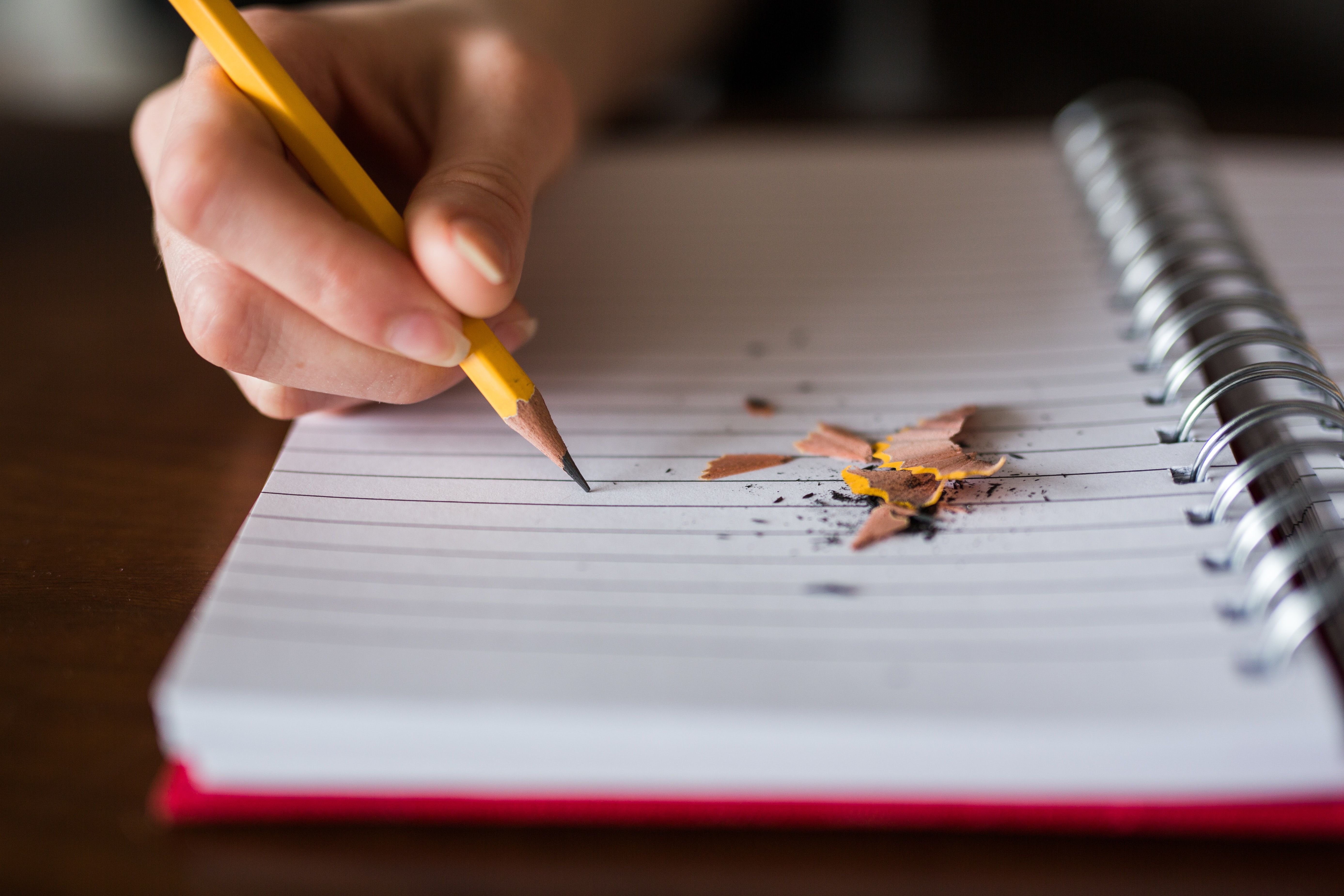 Eine Hand hält einen gelben Bleistift und schreibt auf ein liniertes Blatt, während Bleistiftspäne daneben liegen.