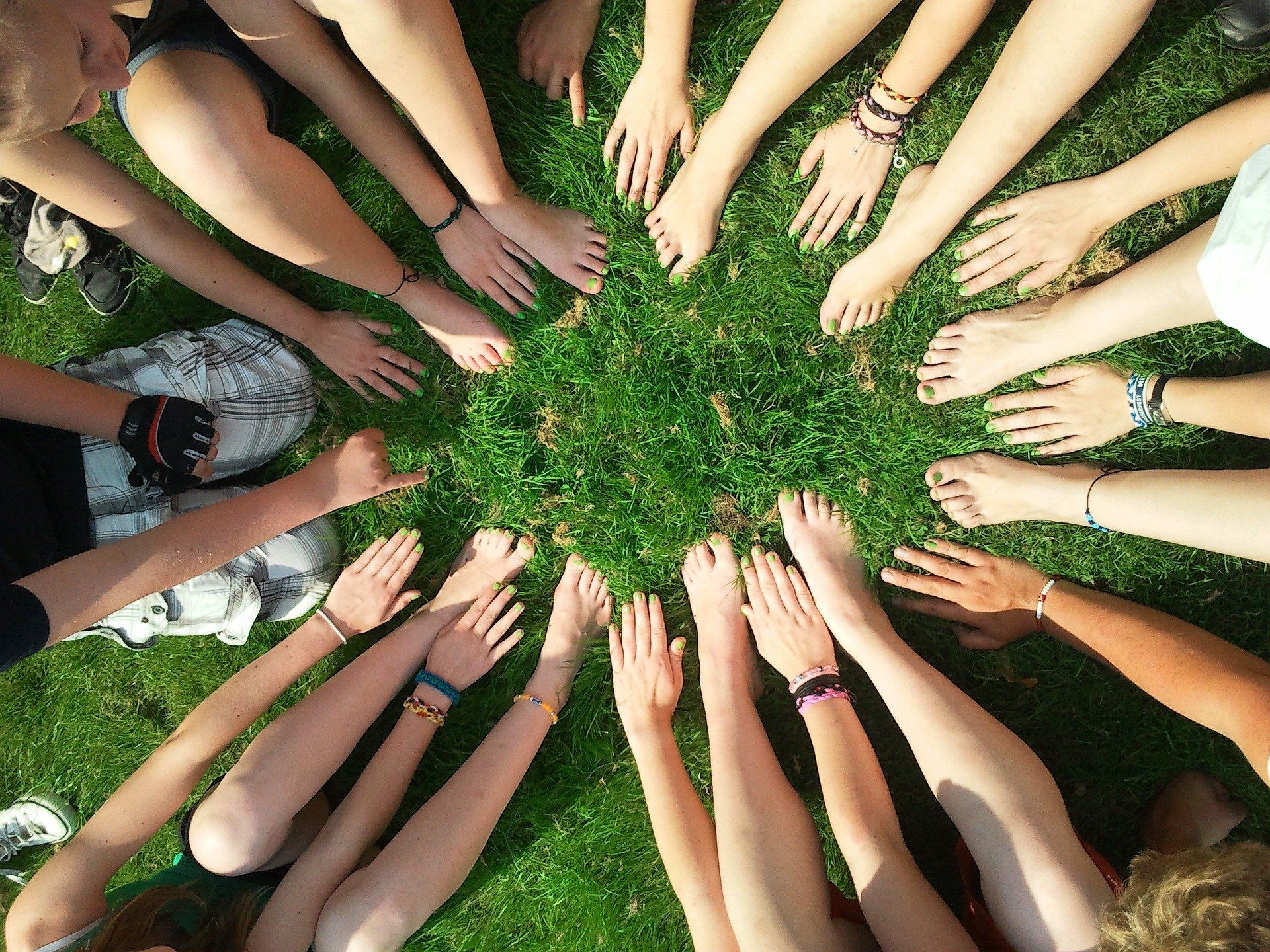 Eine Gruppe von Jugendlichen sitzt im Kreis auf dem Gras, ihre Hände und Füße berühren den Boden, was Gemeinschaft symbolisiert.