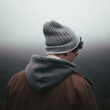 Ein junger Mann mit Mütze und Jacke steht nachdenklich in einer nebligen Landschaft.