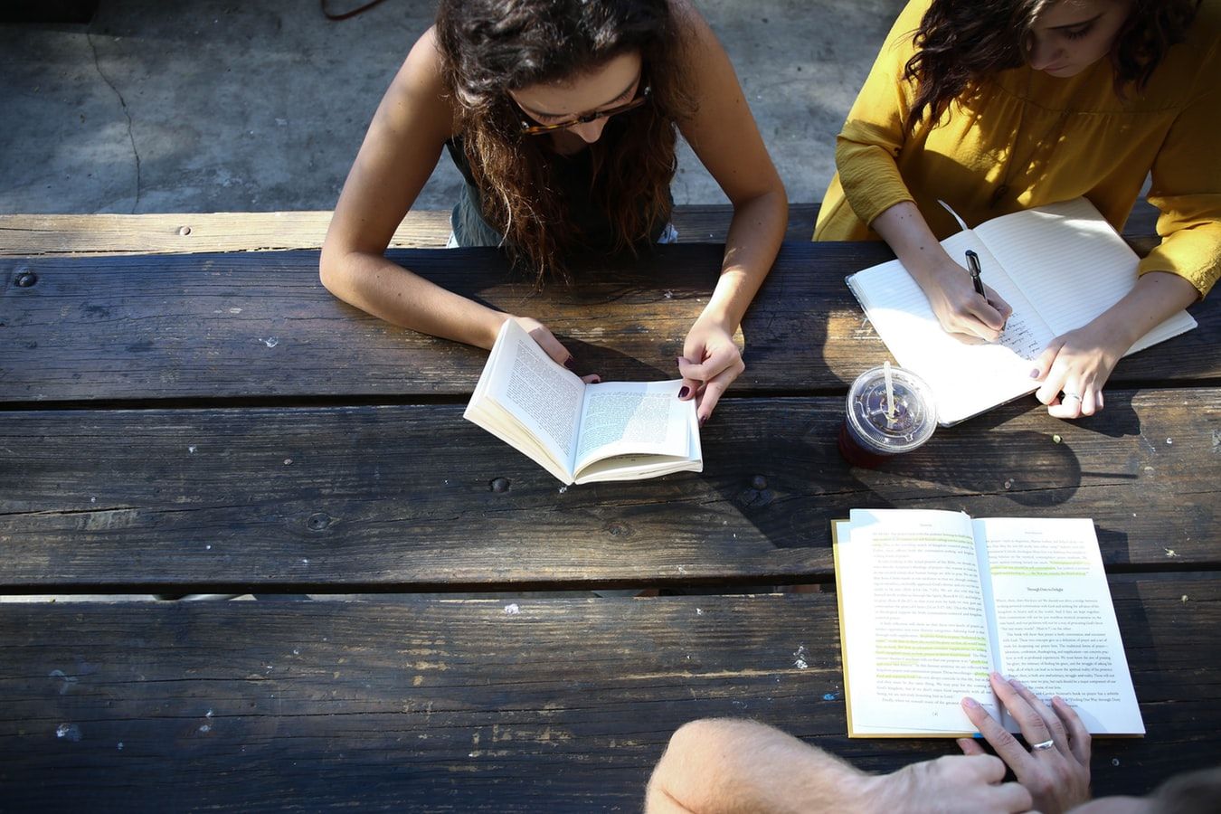 Zwei Personen sitzen an einem Holztisch, lesen Bücher und machen Notizen in einem sonnigen Café.