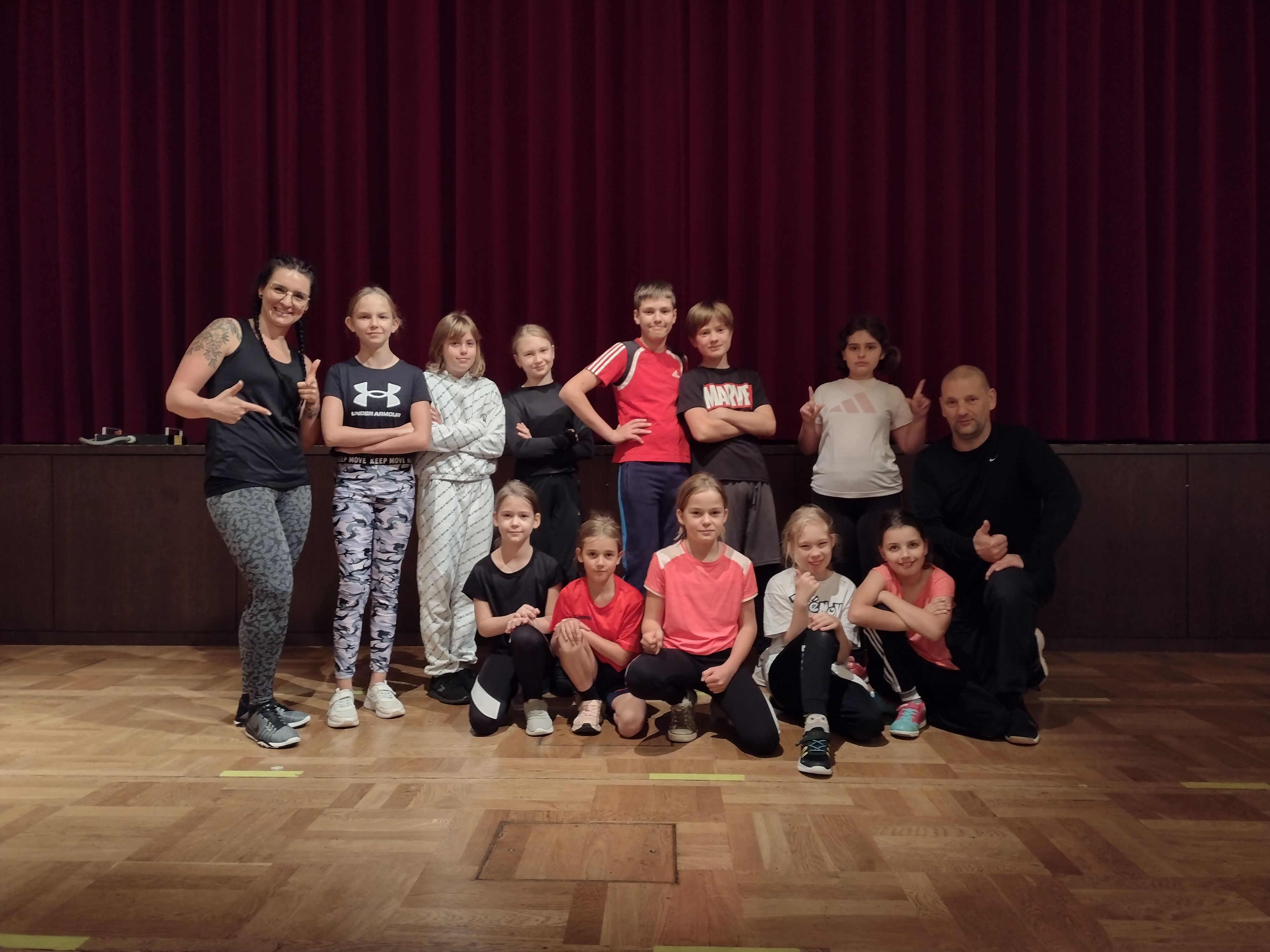 Eine Gruppe von Kindern und Lehrern posiert fröhlich in einem Tanzraum mit rotem Vorhang im Hintergrund.