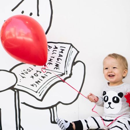 Ein fröhliches Kleinkind sitzt mit einem roten Ballon und einer Spielzeugfigur vor einer Wand mit einer Zeichnung.