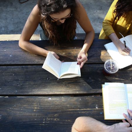 Zwei Personen sitzen an einem Holztisch, lesen Bücher und machen Notizen in einem sonnigen Café.