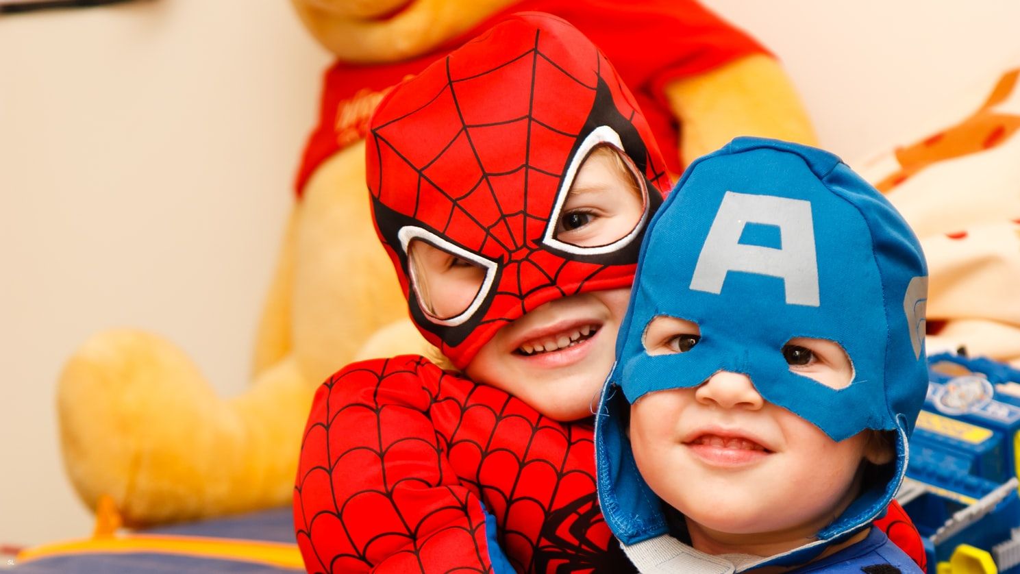 Zwei kleine Kinder in Superheldenkostümen, Spiderman und Captain America, lächeln fröhlich zusammen im Zimmer.
