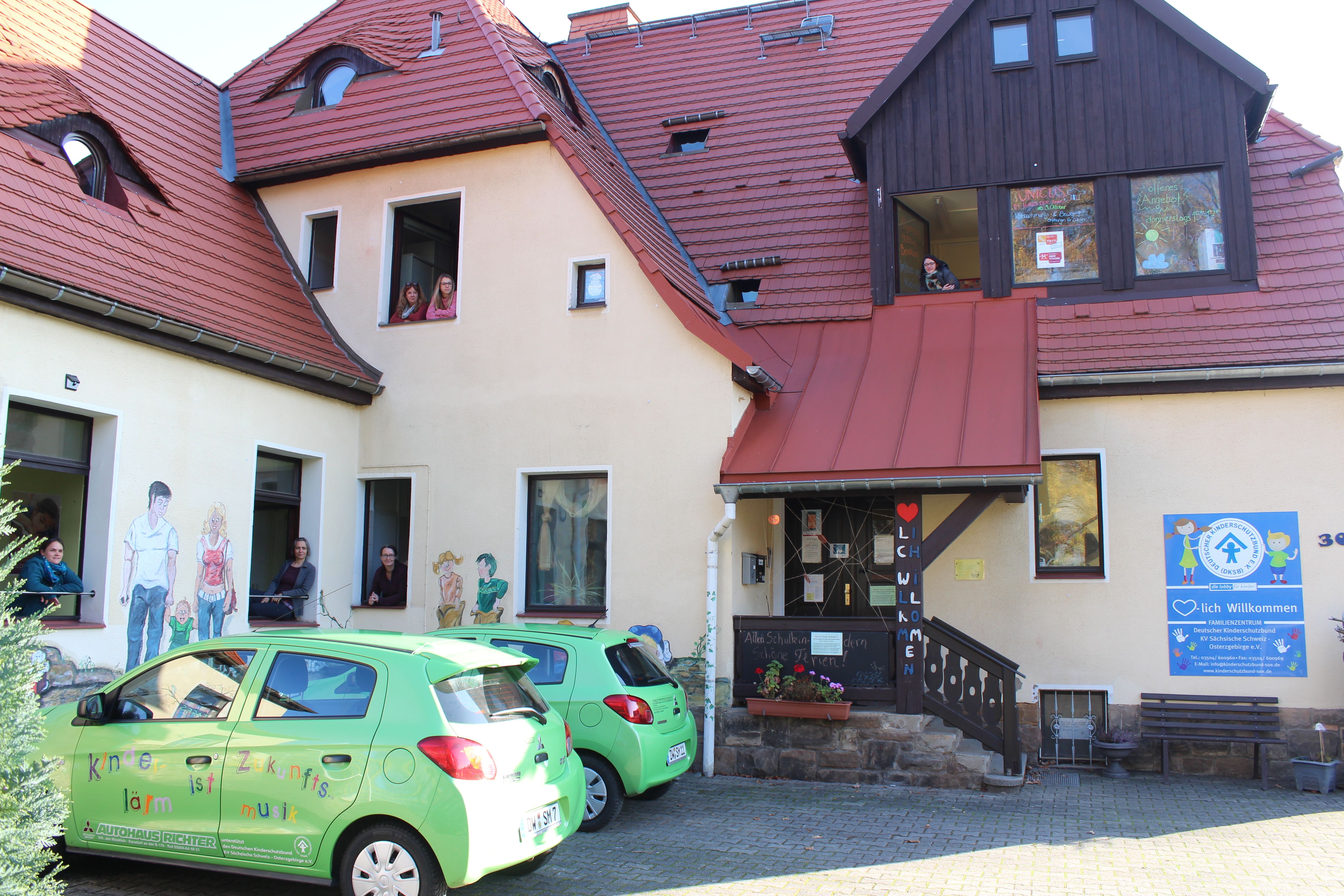 Farbenfrohes Gebäude mit Kindern im Fenster und grünen Autos, die den Willkommen-Bereich der Einrichtung zeigen.