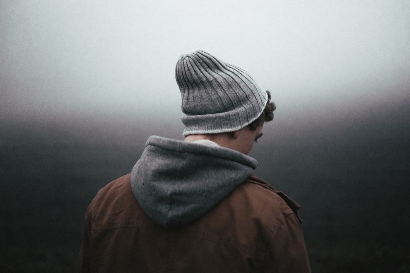Ein junger Mann mit Mütze und Jacke steht nachdenklich in einer nebligen Landschaft.